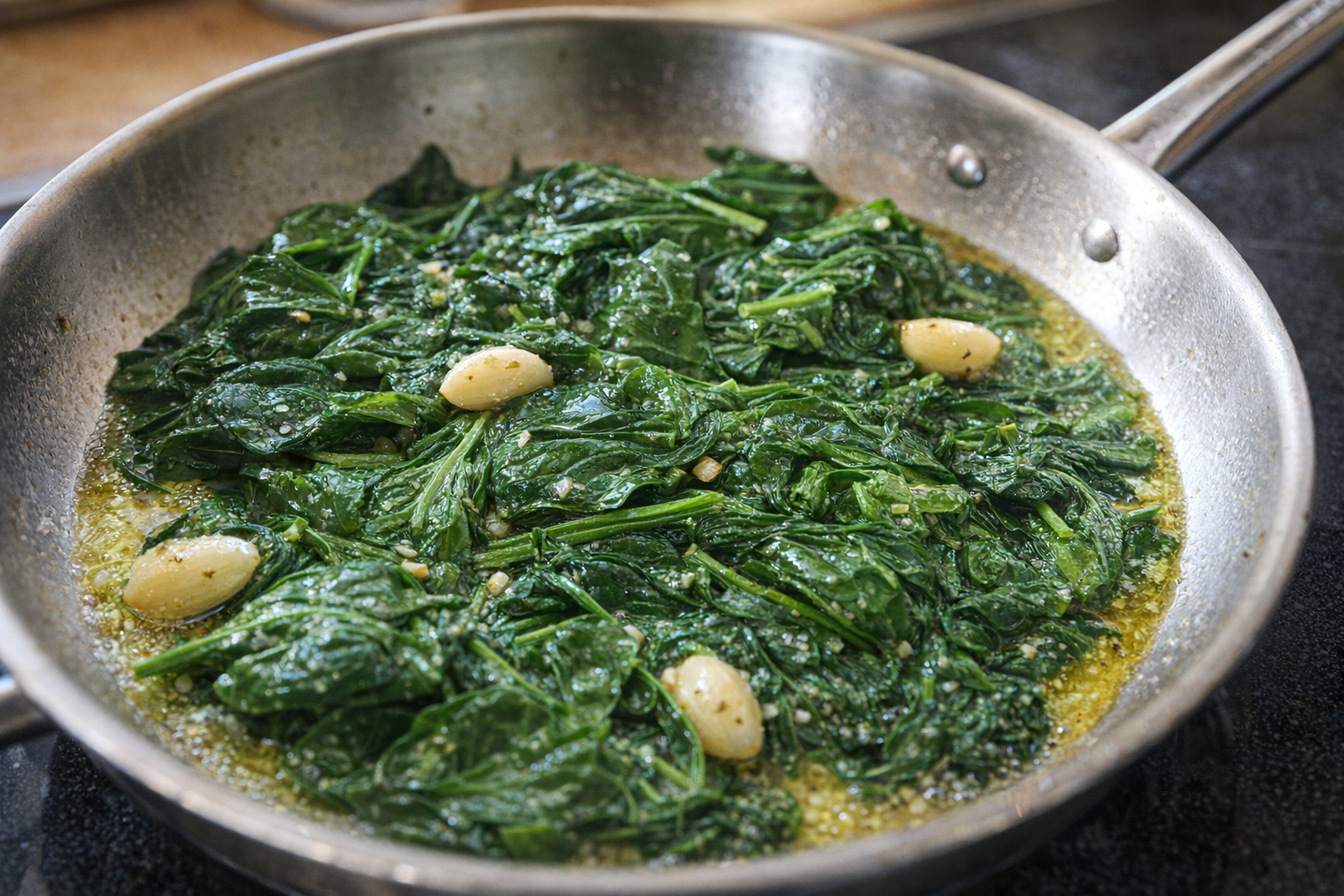 A close-up, overhead shot of vibrant green spinach leaves sautéing in a stainless steel pan with visible garlic cloves, glistening with olive oil under bright kitchen lighting